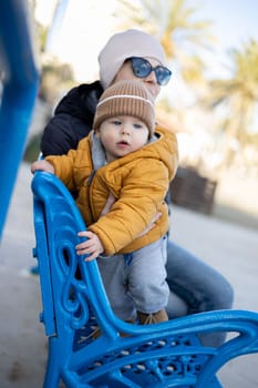 Young mother with her cute infant baby boy child on bench in city park