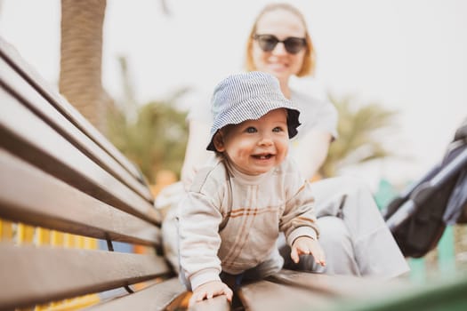 Young mother with her cute infant baby boy child on bench on urban children's playground on warm summer day