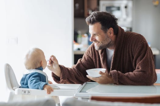 Father wearing bathrope spoon feeding hir infant baby boy child sitting in high chair at the dining table in kitchen at home in the morning.