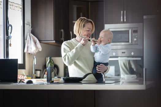 Happy mother and little infant baby boy cooking and tasting healthy dinner in domestic kitchen. Family, lifestyle, domestic life, food, healthy eating and people concept