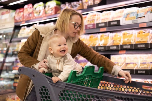 Caucasian mother shopping with her infant baby boy child choosing products in department of supermarket grocery store