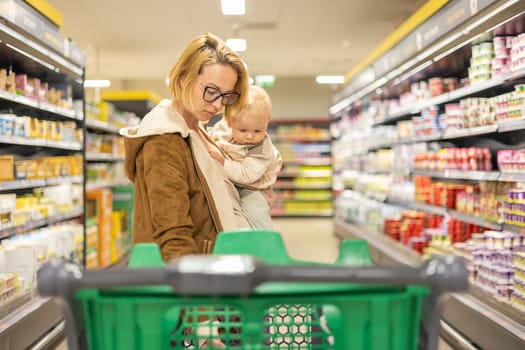 Mother shopping with her infant baby boy child, pushing shopping cart down department aisle in supermarket grocery store