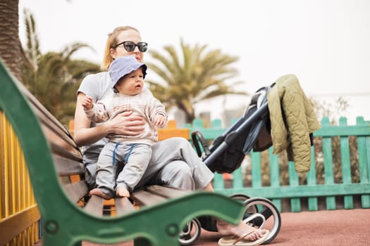 Young mother with her cute infant baby boy child on bench on urban children's playground on warm summer day