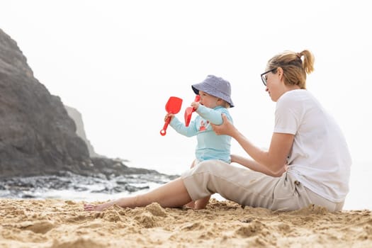 Mother playing his infant baby boy son on sandy beach enjoying summer vacationson on Lanzarote island, Spain. Family travel and vacations concept