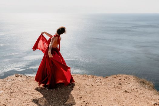 Side view a Young beautiful sensual woman in a red long dress posing on a rock high above the sea during sunrise. Girl on the nature on blue sky background. Fashion photo.