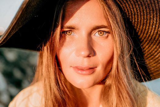 Portrait of happy young woman wearing summer black hat with large brim at beach on sunset. Closeup face of attractive girl with black straw hat. Happy young woman smiling and looking at camera at sea