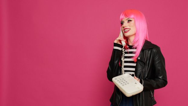 Attractive woman with pink wig talking on landline phone call, chatting on retro telephone line with cord in studio. Having fun using vintage phone for remote communication, cool person.