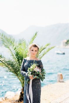Bride with a bouquet of flowers stands on a pier by the sea against the backdrop of mountains. High quality photo