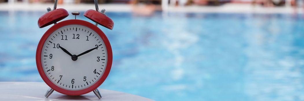 Red alarm clock for ten o'clock near pool in hotel or spa center. Summer vacation tourism and relaxation concept