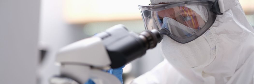 Biologist in protective suit with goggles and medical mask working with microscope in laboratory. Research dangerous biological viruses concept