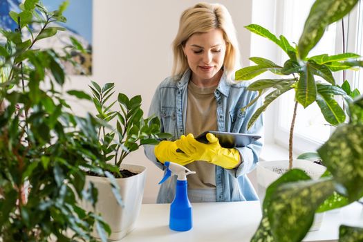 Young happy woman is spraying houseplants with water from a sprayer at home. There are a lot of indoor plants in the room. Home gardening. Hobby concept. Biophilia design, urban jungle concept