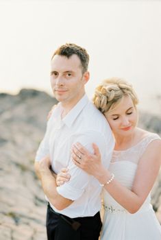 Bride hugs groom from behind with her head resting on his shoulder. Portrait. High quality photo