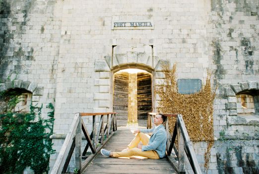 Groom sits on the wooden bridge of Mamula Fort leaning against the railing. High quality photo