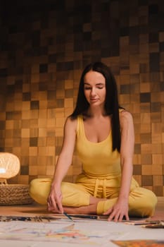 A girl in yellow clothes sitting in the Lotus position draws on paper with evening lighting at home.