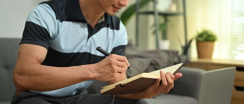 Cropped image of man freelancer sitting on couch and making important notes, planning daily appointment on notepad.