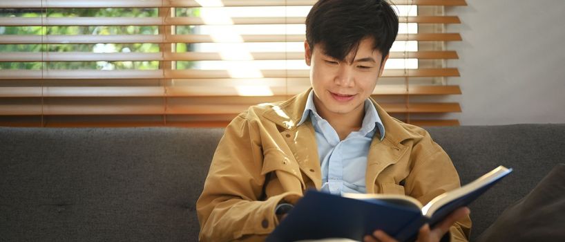 Pleased asian man sitting on comfortable sofa and reading interesting book, enjoying leisure weekend time at home.