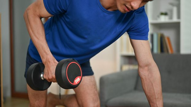 Muscular asian man bodybuilder exercising with heavy dumbbell at home. Healthy lifestyle and fitness concept.