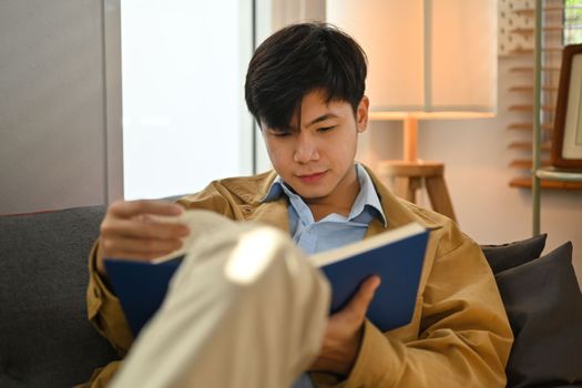 Peaceful millennial man reading interesting book on comfortable sofa, enjoying leisure weekend time at home.