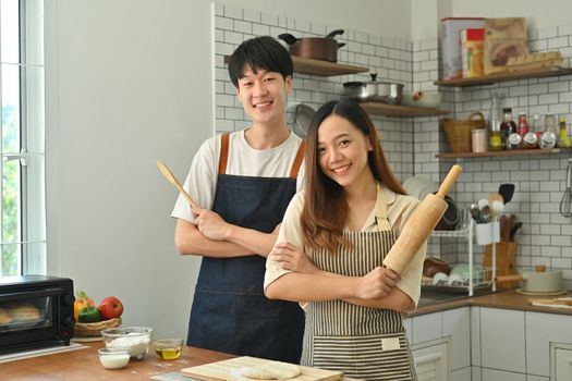 Happy married couple with rolling pins in their hand, preparing dinner in modern kitchen, enjoying leisure time at home.