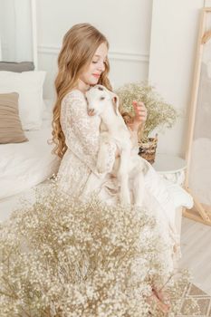 A blonde girl with long hair is sitting on the couch with a goat at home. The concept of animal care