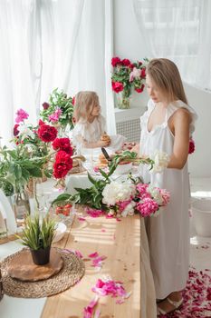 A little blonde girl with her mom on a kitchen countertop decorated with peonies. The concept of the relationship between mother and daughter. Spring atmosphere.