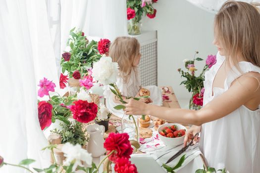 A little blonde girl with her mom on a kitchen countertop decorated with peonies. The concept of the relationship between mother and daughter. Spring atmosphere.