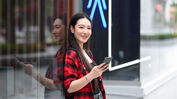 Cheerful Asian woman standing on city street and using smart phone.