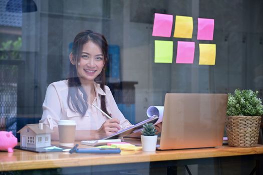 View through glass window young businesswoman sitting in workplace and smiling to camera.