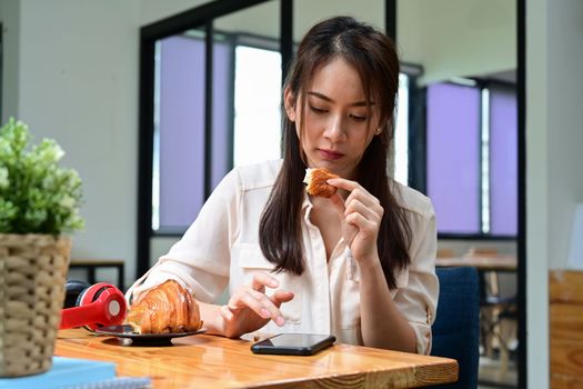 Young female having breakfast and using smart phone at her workplace.