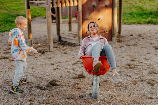 Mother and son having fun on a swing. Motherhood and childhood concept. Sunny summer day. Beautiful family is having fun outside. Parents with children riding on a swing. Mom is playing with her little son on a terrace with swings. A mother woman swings on a swing in the park. Mom and child are on a swing in nature