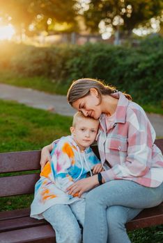 mother and son sit on a park bench in the rays of the setting sun. the concept of a family. Mother's Day. beautiful girl (mother) with a boy (son) in the park in the park are sitting on a bench at sunset.