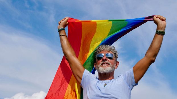 Portrait of a gray-haired elderly Caucasian man with a beard and sunglasses holding a rainbow LGBTQIA flag against a sky background. Bisexual Gay Celebrates Pride Month Coming Out Day