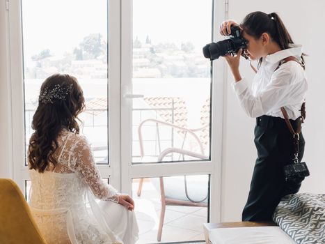 Girl photographer photographs the bride sitting on a chair near the balcony door. High quality photo