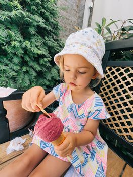 Little girl in a panama hat sits on a bench and eats a cone of popsicles with a spoon. High quality photo