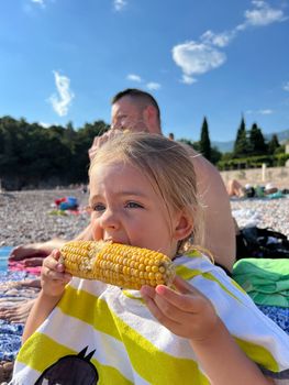 Little girl eating a head of corn while sitting on the beach. High quality photo