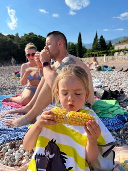 Man, woman and little girl are sitting on the beach and eating corn. High quality photo