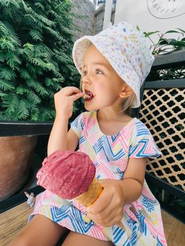 Little girl sits on a bench near a tree and eats a popsicle cone with a spatula. High quality photo