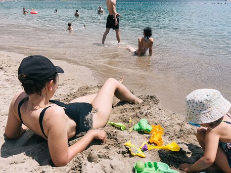 Mom sunbathing on the beach next to a little girl sculpting sand figures. High quality photo