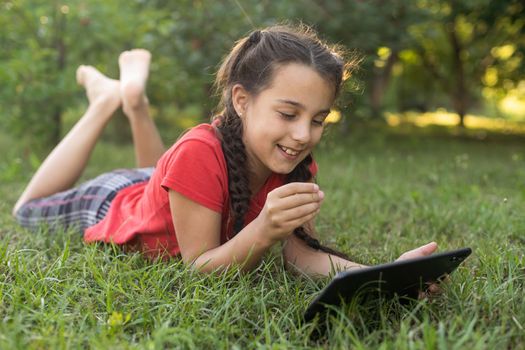 Distance learning during the quarantine period for the virus Covid-19. Cute little schoolgirl with long hair is studying from home, sitting in the garden on the grass. Uses a tablet and remote work