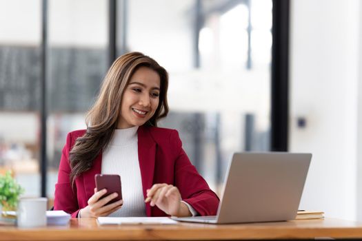 Smiling beautiful Asian businesswoman analyzing chart and graph showing changes on the market and holding smartphone at office.