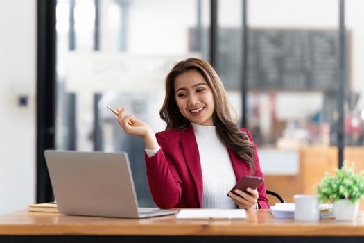 Smiling beautiful Asian businesswoman analyzing chart and graph showing changes on the market and holding smartphone at office.