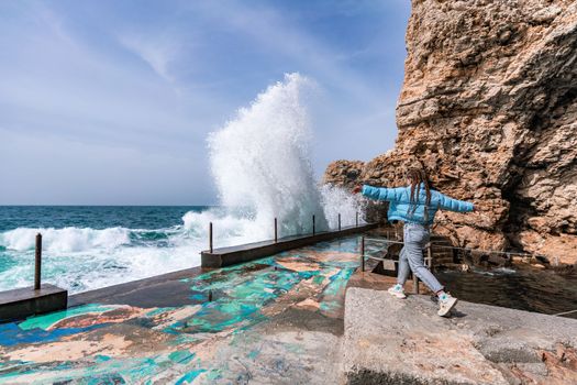 A woman in a blue jacket stands on a rock above a cliff above the sea and looks at the raging ocean. Girl traveler rests, thinks, dreams, enjoys nature. Peace and calm landscape, windy weather