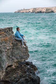 Woman rocks sea. A woman in a blue jacket sits on a rock above a rock above the sea and looks at the raging ocean. Girl traveler rests, thinks, dreams, enjoys nature