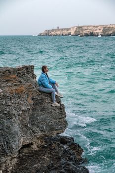 A woman in a blue jacket sits on a rock above a cliff above the sea, looking at the stormy ocean. Girl traveler rests, thinks, dreams, enjoys nature. Peace and calm landscape, windy weather