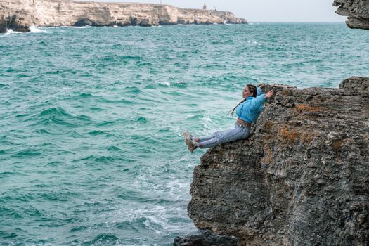 Woman rocks sea. A woman in a blue jacket sits on a rock above a rock above the sea and looks at the raging ocean. Girl traveler rests, thinks, dreams, enjoys nature