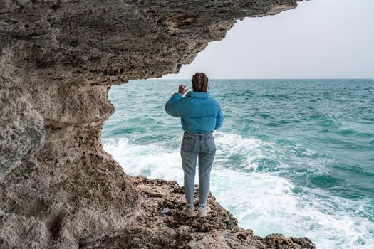 A woman in a blue jacket stands on a rock above a cliff above the sea and looks at the raging ocean. Girl traveler rests, thinks, dreams, enjoys nature. Peace and calm landscape, windy weather