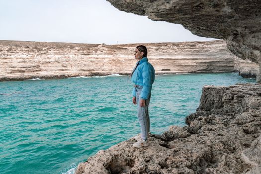 A woman in a blue jacket stands on a rock above a cliff above the sea and looks at the raging ocean. Girl traveler rests, thinks, dreams, enjoys nature. Peace and calm landscape, windy weather