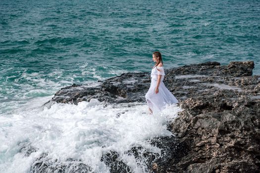 A woman stands on a rock in the sea during a storm. Dressed in a white long dress, the waves break on the rocks and white spray rises