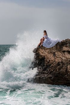 A woman in a storm sits on a stone in the sea. Dressed in a white long dress, waves crash against the rocks and white spray rises above her