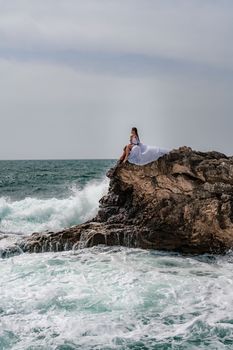 A woman in a storm sits on a stone in the sea. Dressed in a white long dress, waves crash against the rocks and white spray rises